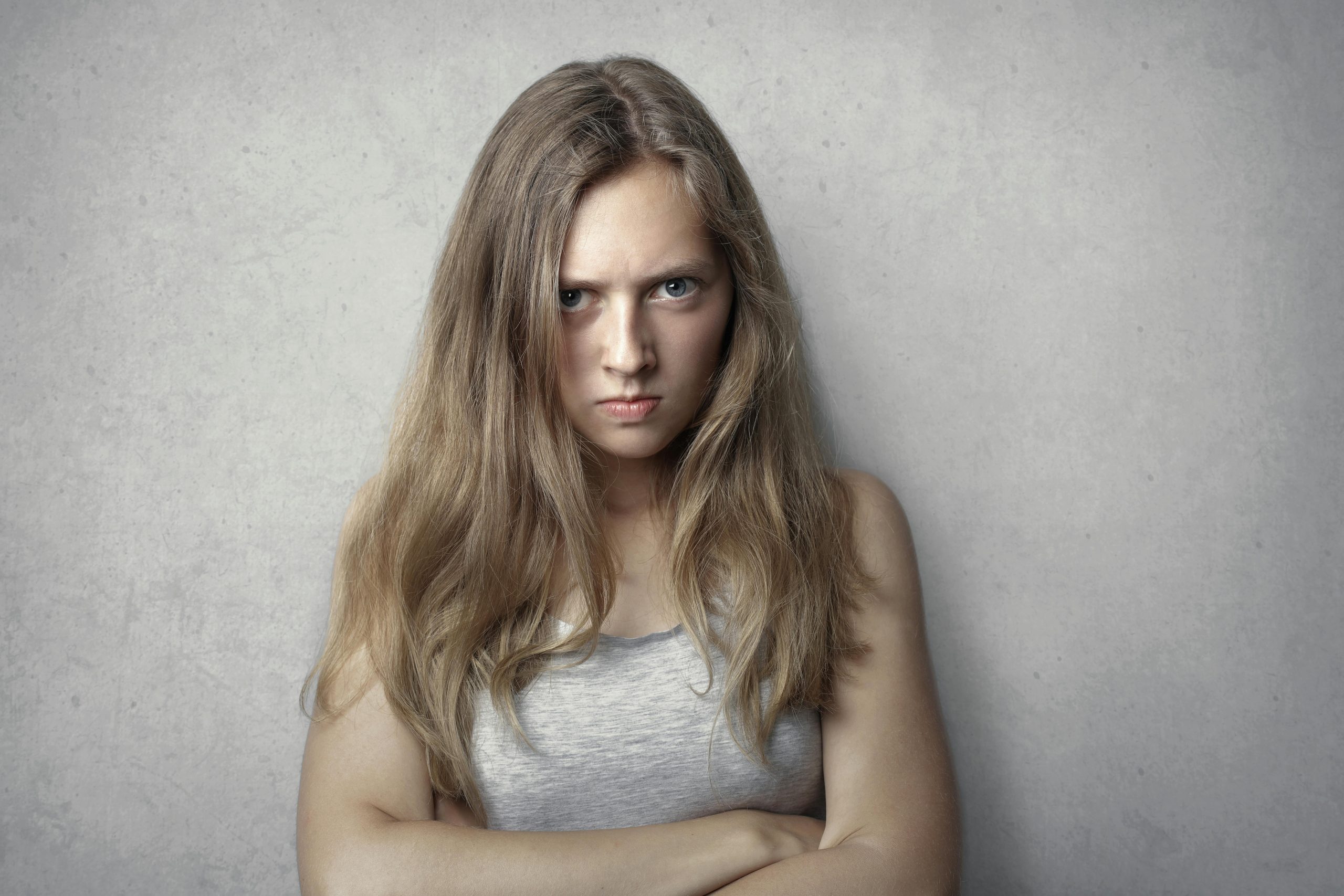 Portrait d'une femme déterminée aux bras croisés avec une expression intense sur fond gris.