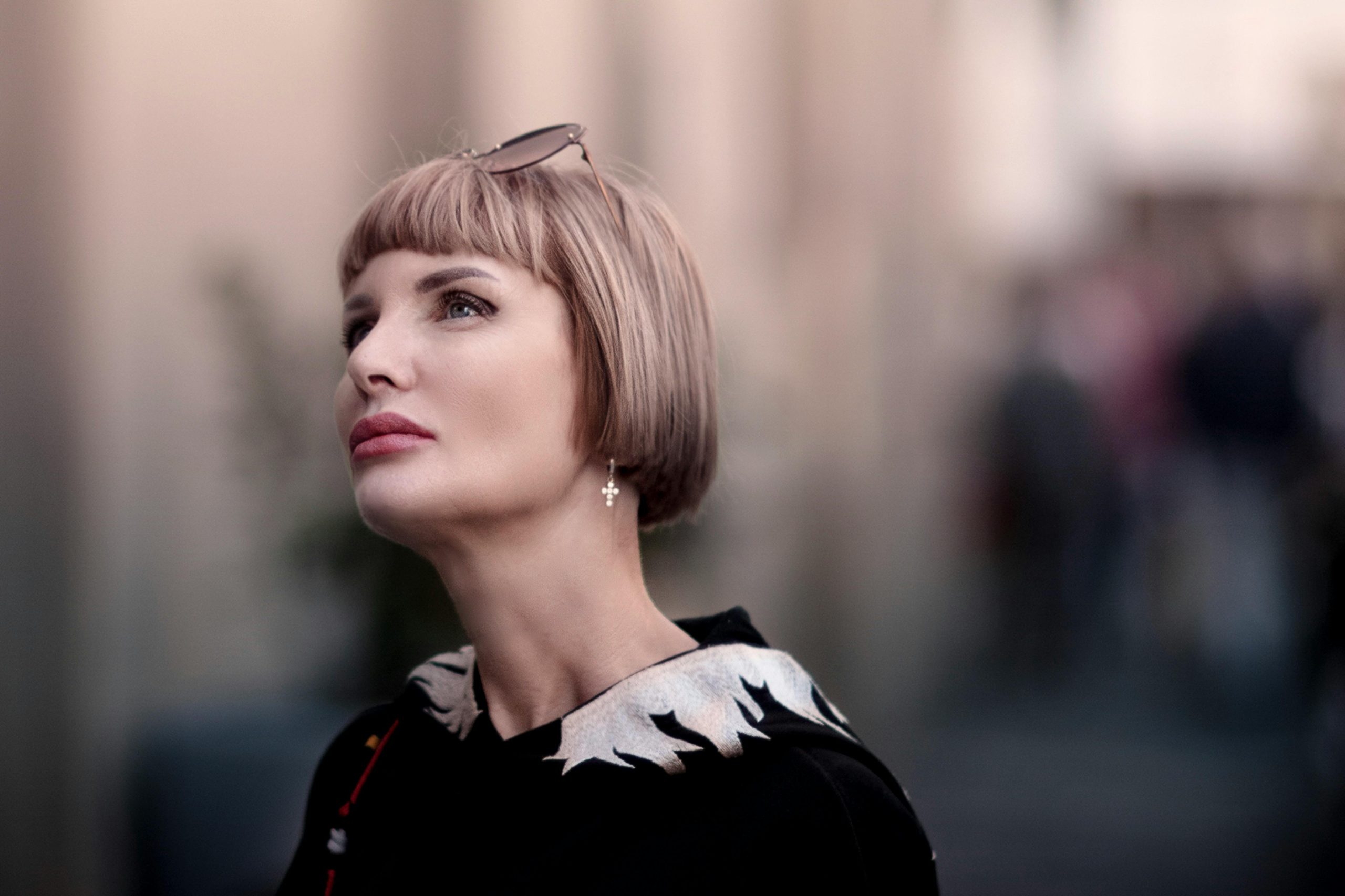 Stylish portrait of a woman with blonde bob haircut and sunglasses, looking up with a blurred urban background. Portrait élégant d'une femme avec une coupe au carré blonde et des lunettes de soleil, regardant vers le haut avec un arrière-plan urbain flou.