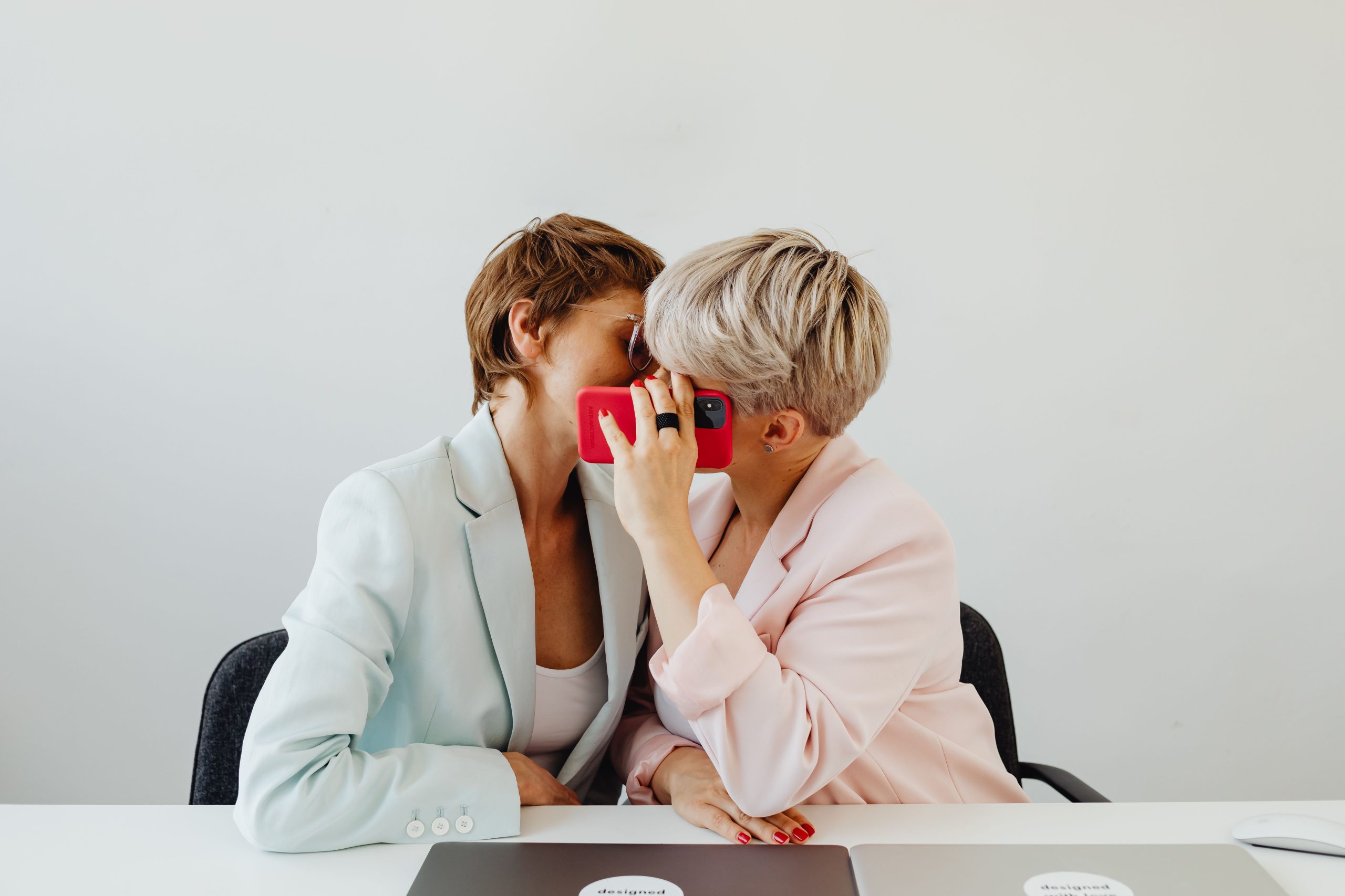 Two women share a secretive moment at a desk in a modern office setting. Deux femmes partagent un moment secret à un bureau dans un cadre de bureau moderne.