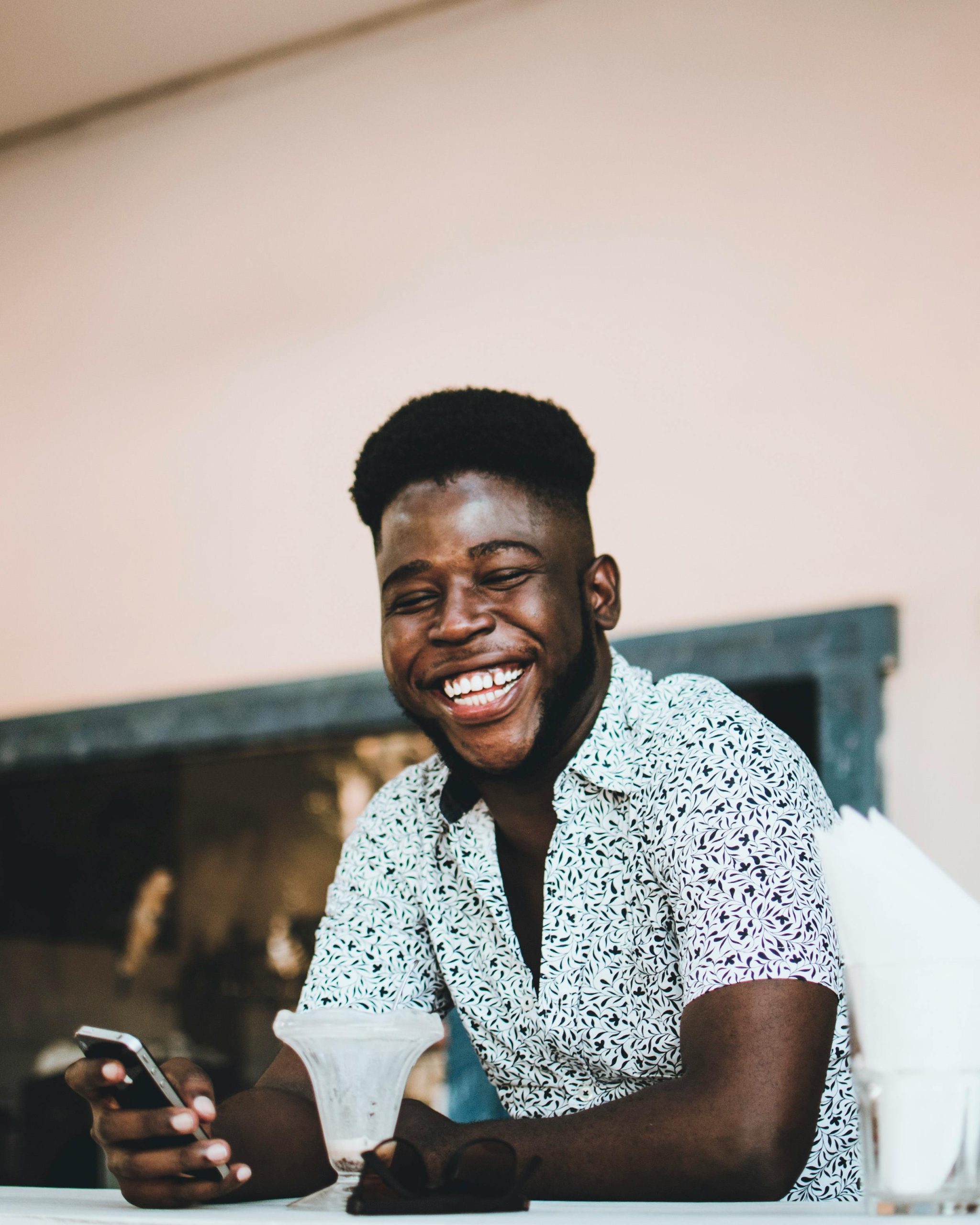 Happy African American man smiling while holding a phone and enjoying coffee indoors. Homme afro-américain heureux souriant en tenant un téléphone et savourant un café à l'intérieur.