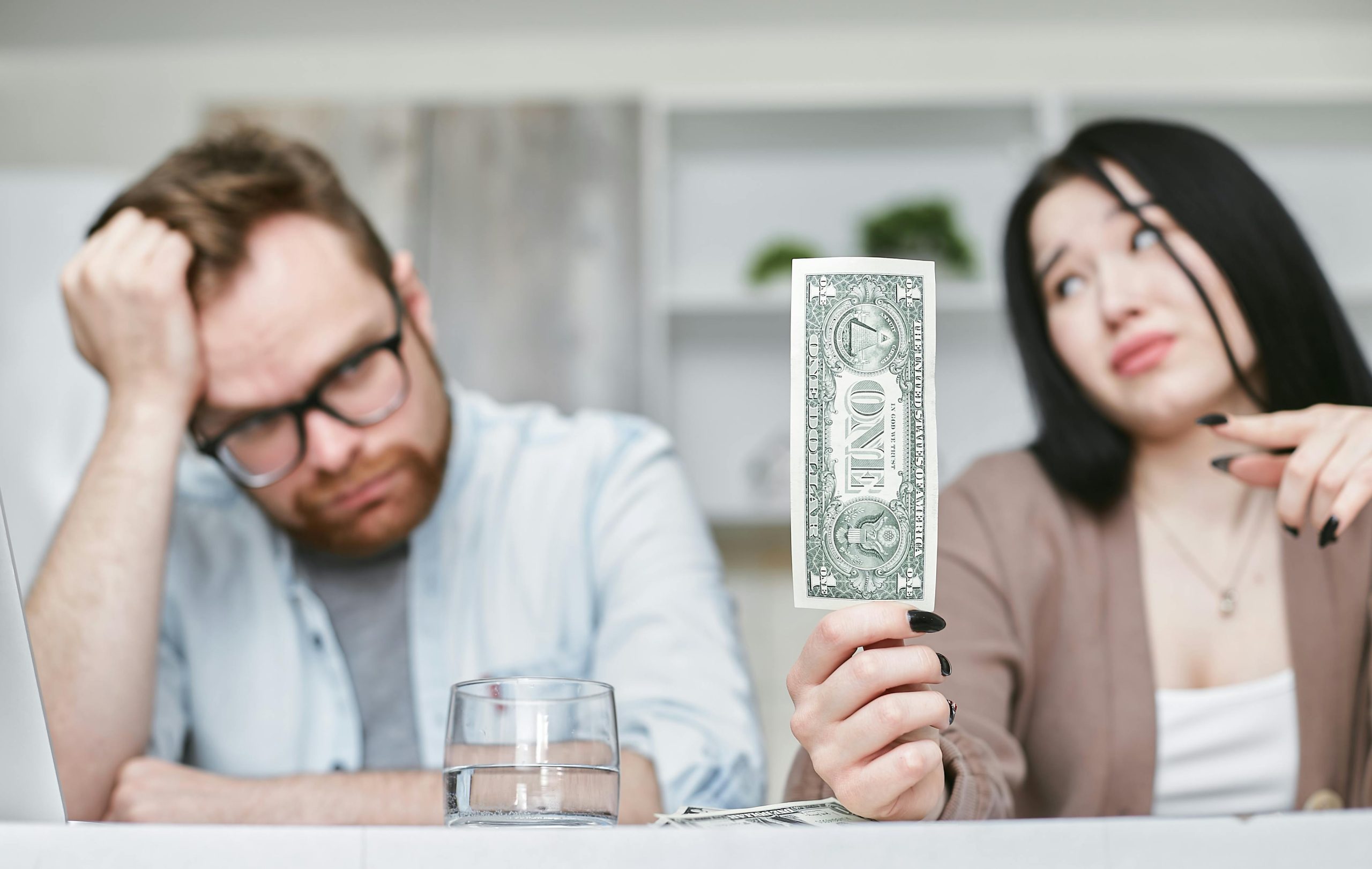 An adult interracial couple expressing concern about finances as they hold a one dollar bill indoors. Un couple interracial adulte exprimant des préoccupations concernant les finances en tenant un billet d'un dollar à l'intérieur.