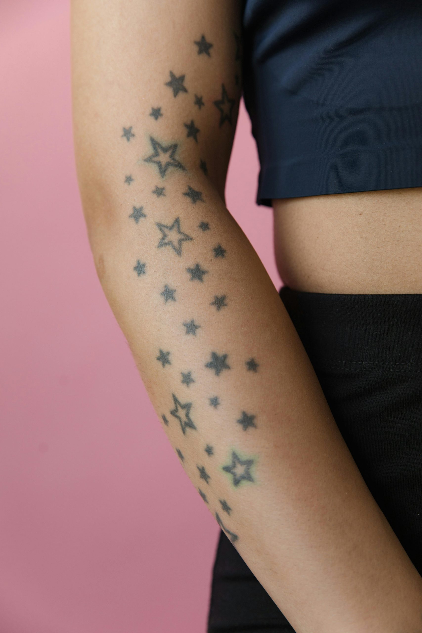 A close-up photo of an arm with star tattoos against a pink backdrop. Une photo en gros plan d'un bras avec des tatouages d'étoiles sur un fond rose.
