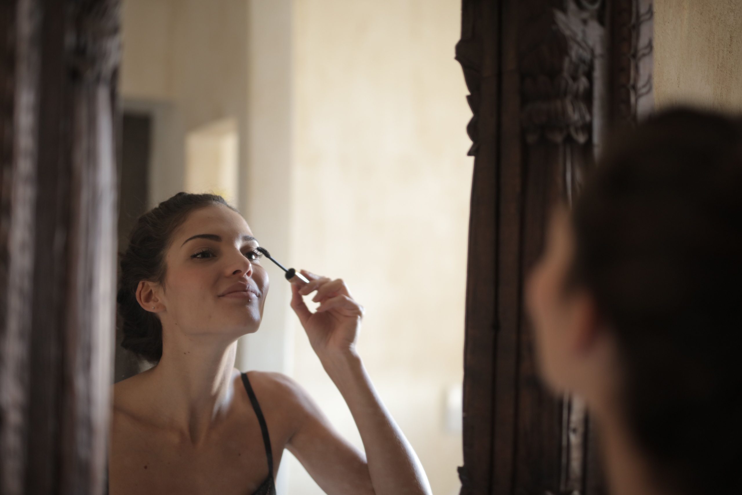 A woman applies mascara while looking in a stylish wooden mirror indoors. Une femme applique du mascara en se regardant dans un miroir en bois élégant à l'intérieur.