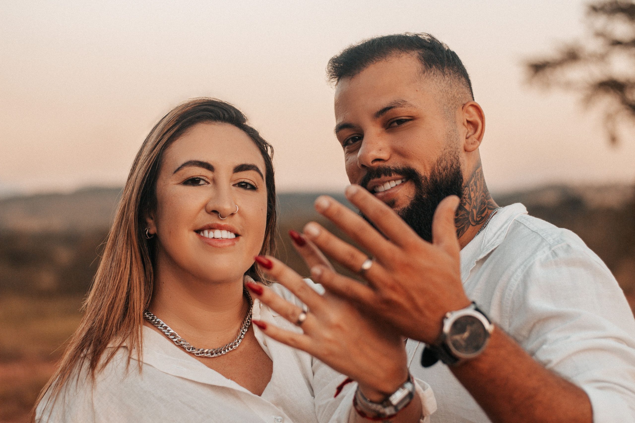 Smiling couple joyfully displaying their engagement rings in an outdoor setting. Un couple souriant affichant joyeusement ses bagues de fiançailles dans un cadre extérieur.