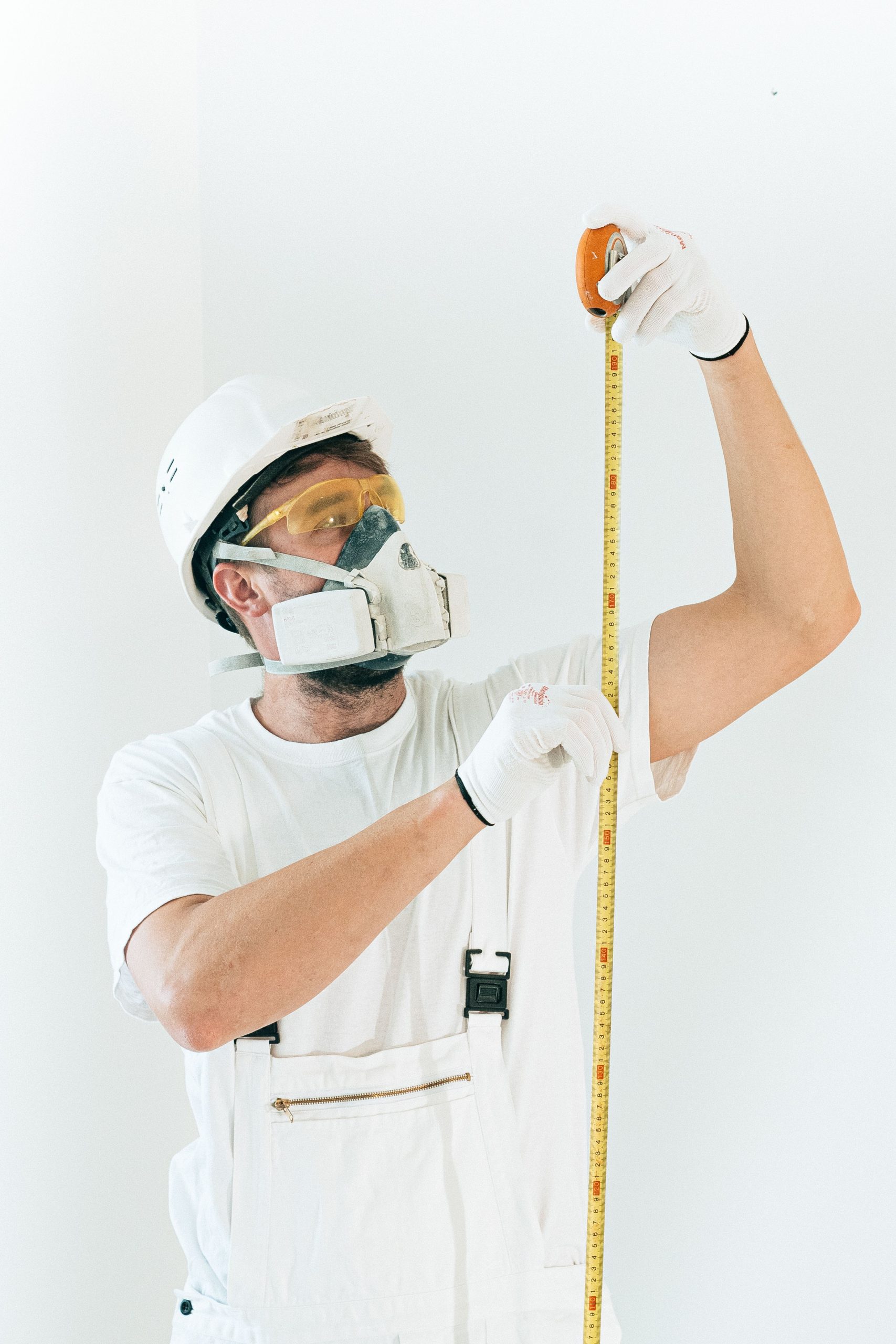 A construction worker in full safety gear measures with a tape against a white wall. Un ouvrier de la construction en équipement de sécurité complet mesure avec un mètre ruban contre un mur blanc.