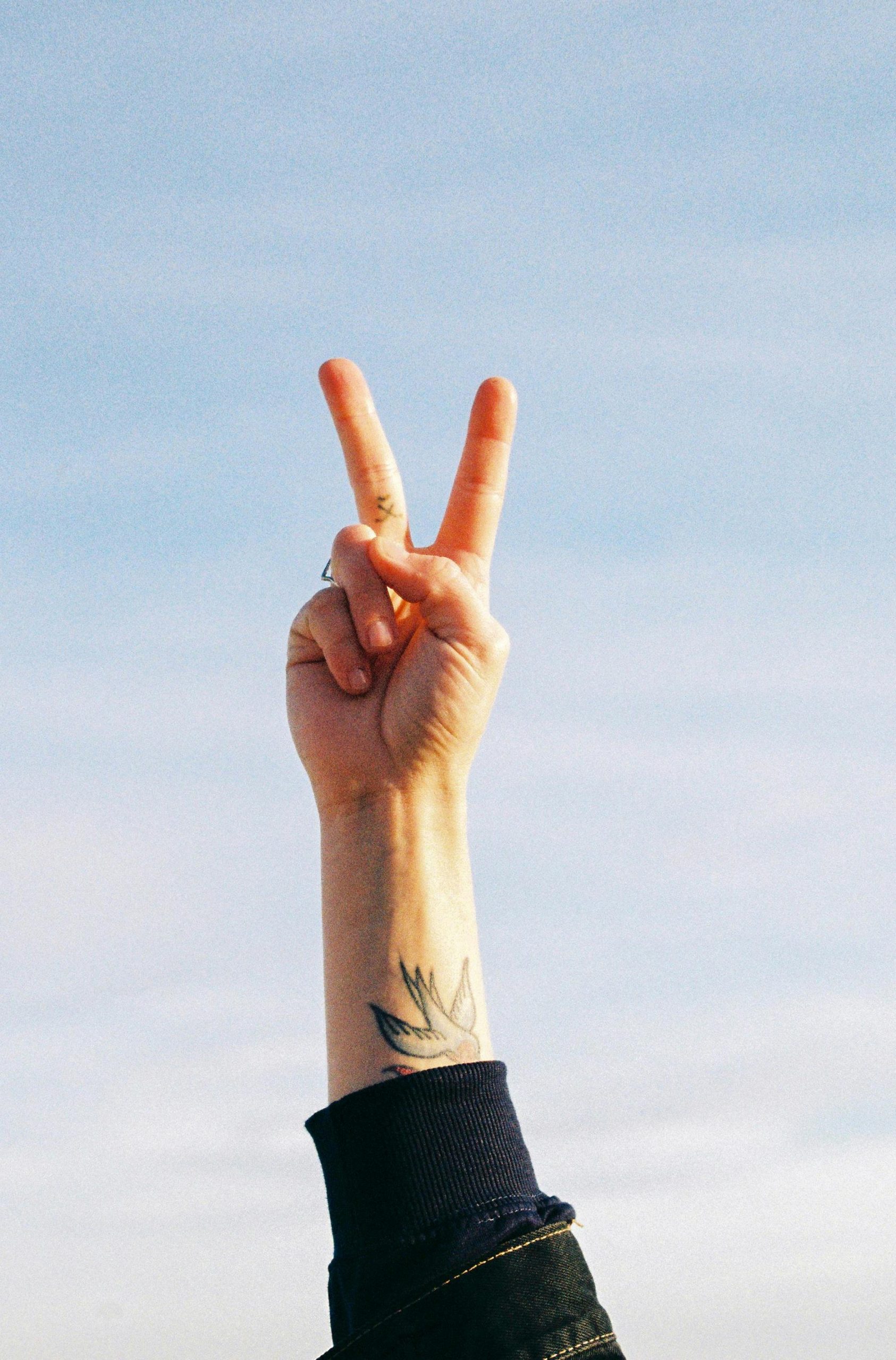 An arm with a tattoo displays a peace sign against a clear blue sky, symbolizing victory and success. Un bras tatoué affiche un signe de paix contre un ciel bleu clair, symbolisant la victoire et le succès.