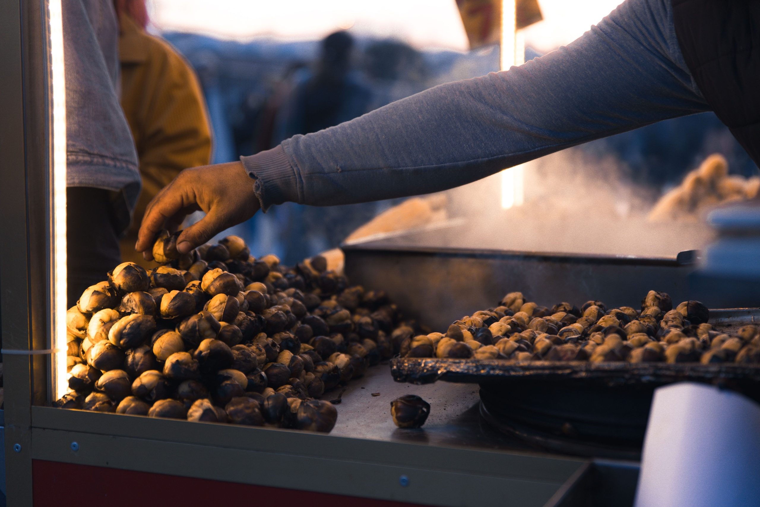 A street vendor's hand reaches over a pile of roasted chestnuts at an outdoor market stall at night. La main d'un vendeur ambulant s'étend sur un tas de châtaignes rôties à un étal du marché en plein air la nuit.