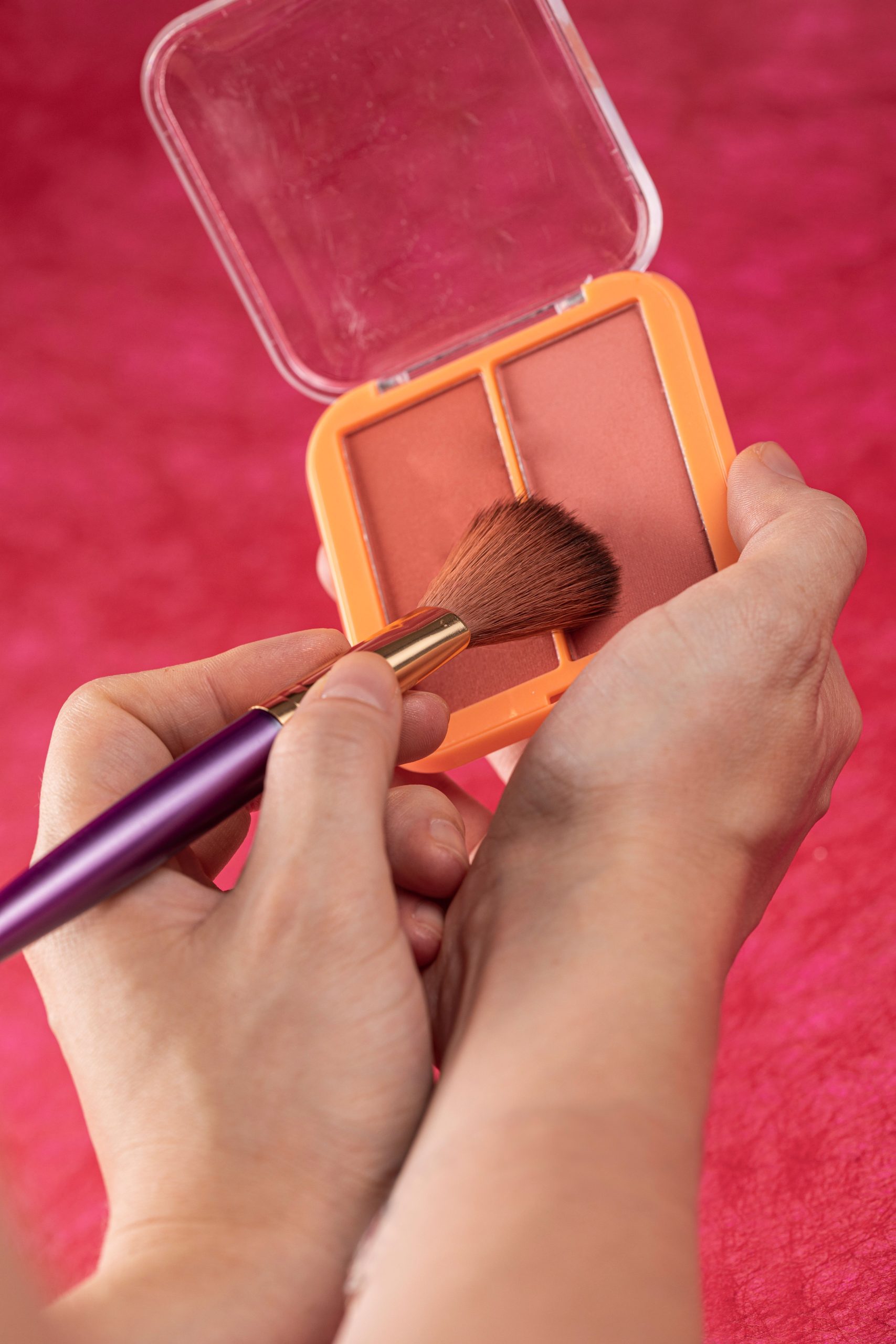 Hands applying blush with a makeup brush on a red background. Les mains appliquent du blush avec un pinceau de maquillage sur un fond rouge.