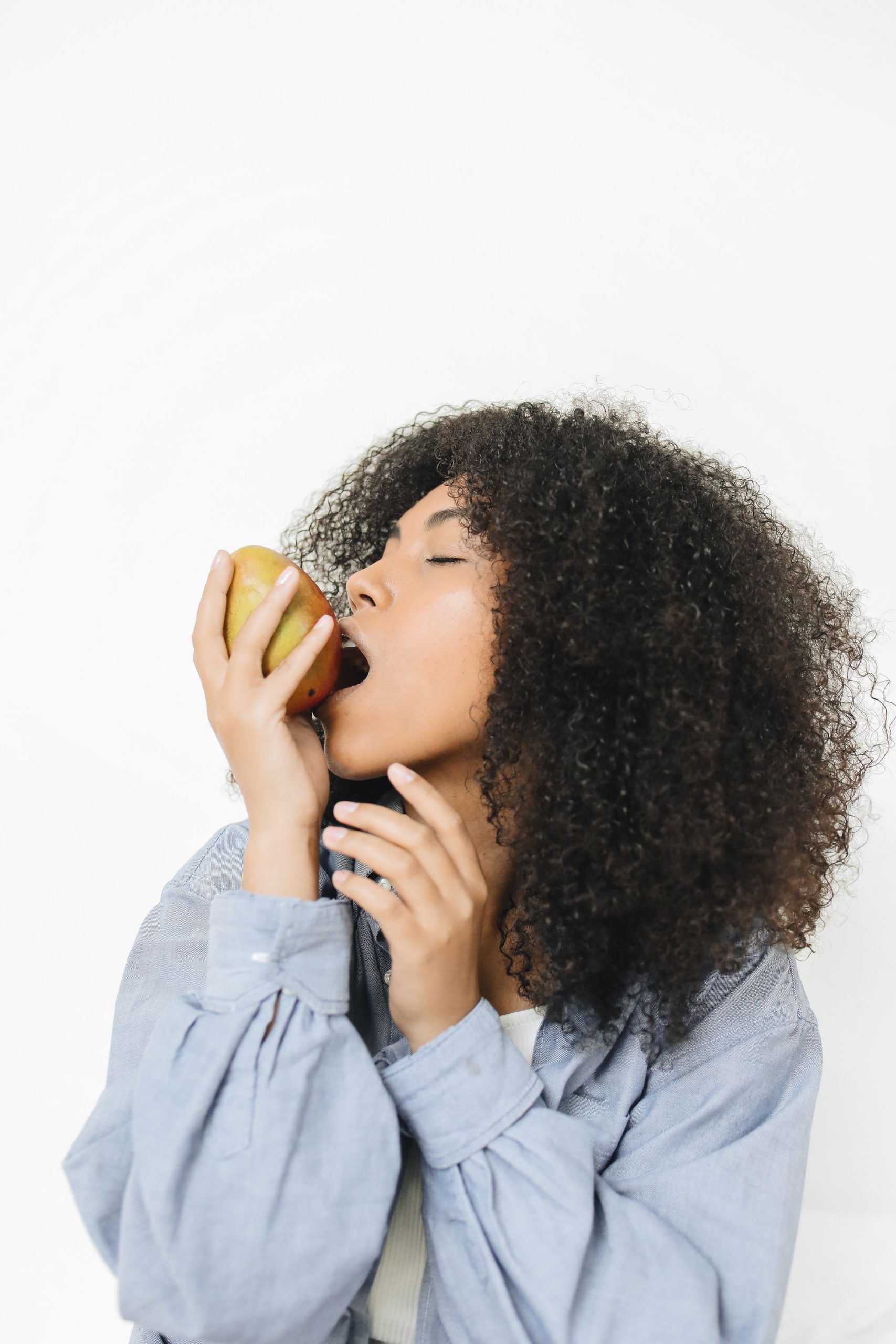 African American woman in studio savoring a healthy, organic mango. White background. Femme afro-américaine en studio savourant une mangue saine et biologique. Fond blanc.
