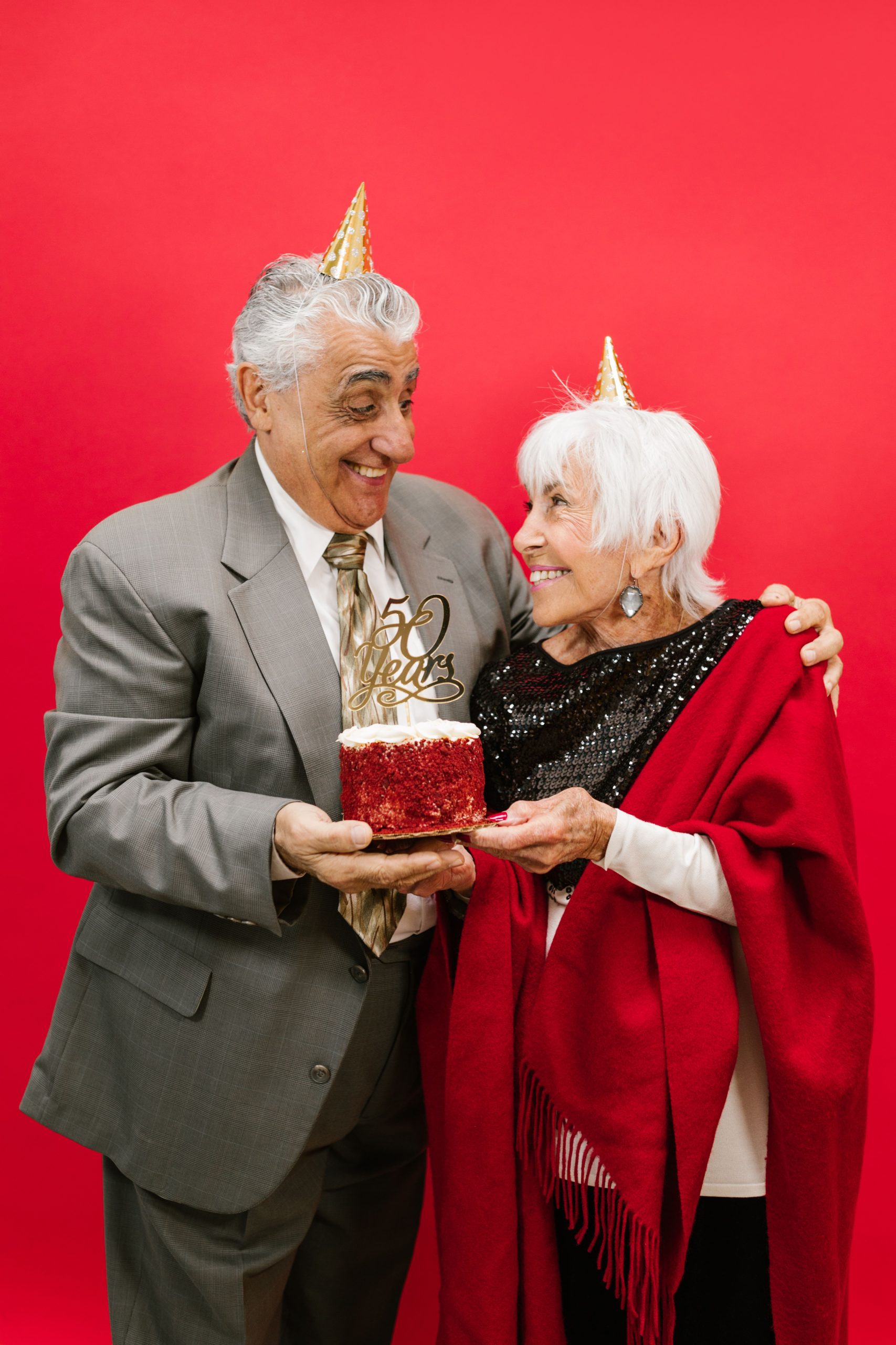 Happy elderly couple celebrating their 50th anniversary with a red cake against a vibrant background. Un couple âgé heureux célébrant son 50e anniversaire avec un gâteau rouge sur un fond vibrant.
